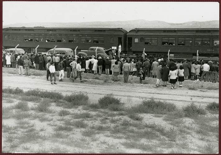 A crowd of people stands on one side of a barbed wire fence. Four cars and a train are on the other side of the fence.
