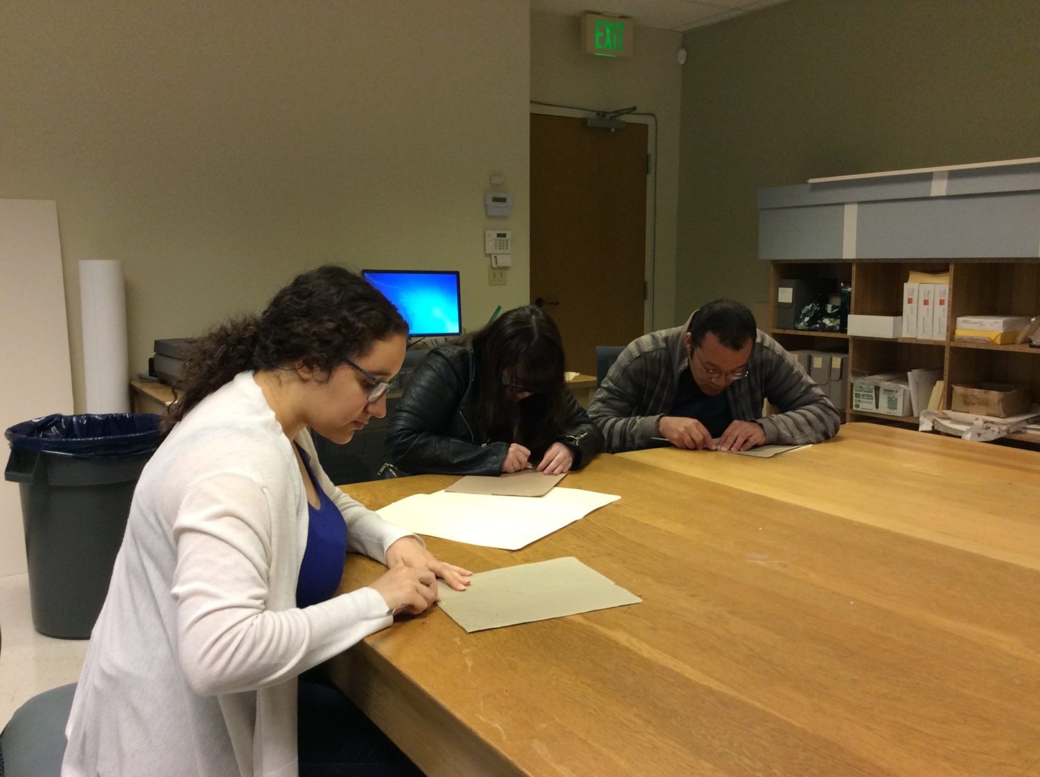 Three students examine documents at a large wooden table.