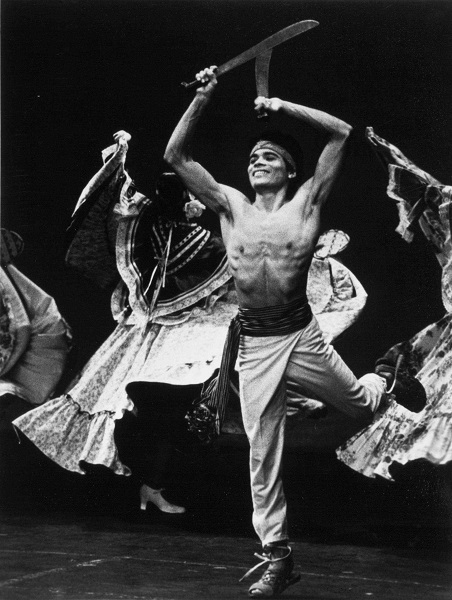 Black and white photo of male dancer in foreground and female dancers in background. Male dancer is shirtless and holding two crossed sabers above his head.
