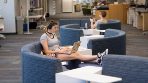 A student sitting in round seating in an open study space in the library. One student in on her laptop wearing headphones and another student in the background in sitting in a chair, texting on his cell phone.