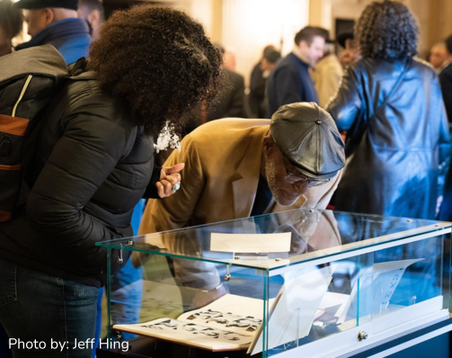 Decorative image showing two people standing over a glass exhibit box peering in to look closer at a book on display.