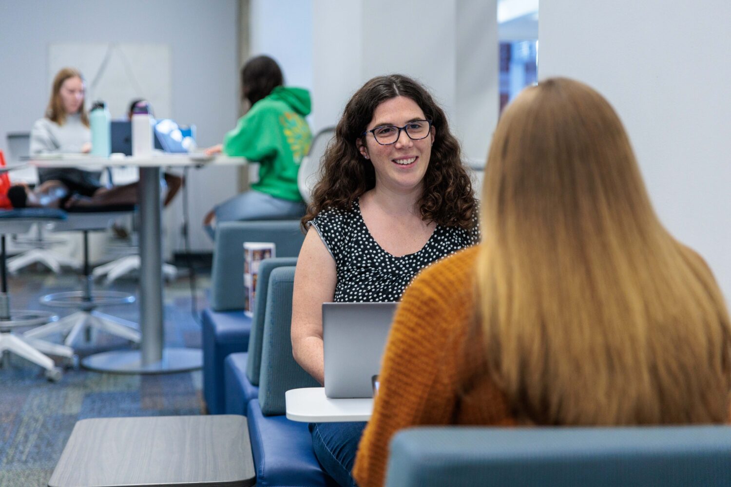 A smiling woman with curly dark hair and glasses sits at a study lounge table in The Claremont Colleges Library, speaking with another person in the foreground. Both have laptops open, and other students work at high-top tables in the background, creating a collaborative academic atmosphere.