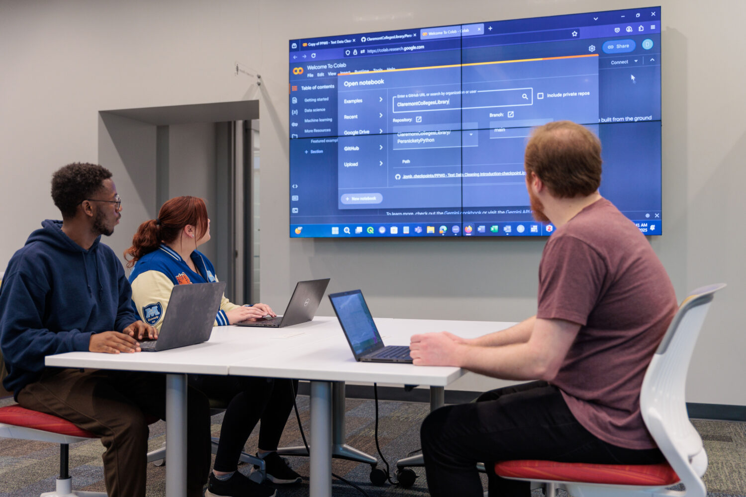 Three students sit at a table in The Claremont Colleges Library, each with a laptop open, looking at a large wall-mounted screen displaying a coding notebook interface. They appear to be discussing or learning data analysis together in a collaborative workspace.