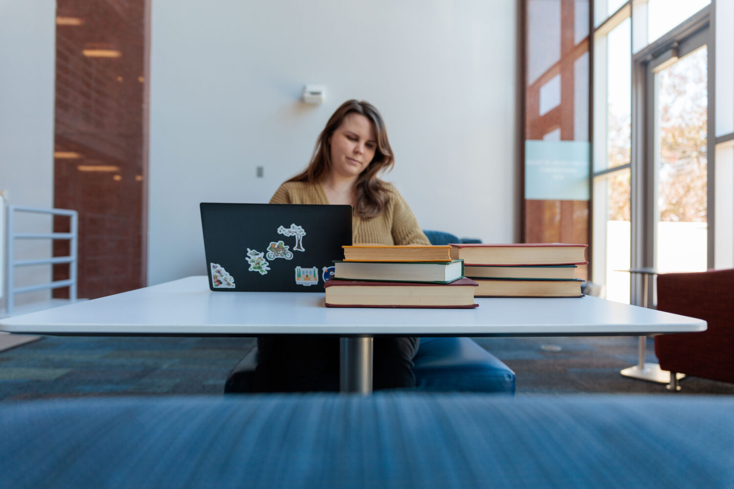 A woman works on a laptop covered in colorful stickers at a white table in a bright, modern seating area of The Claremont Colleges Library, with several stacks of books beside her.